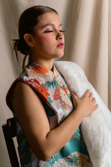 A young girl with closed eyes and a serene expression, wearing a colorful, floral-patterned kimono-style garment, gently holding a soft white blanket close to her chest. She has dark brown hair pulled